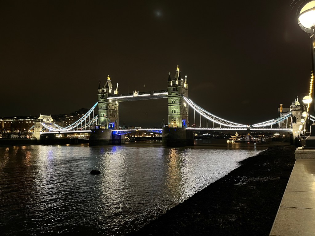 Tower Bridge lit up at night, with its 2 tall ornate towers linked by the road between them below and a footbridge between them near the top.