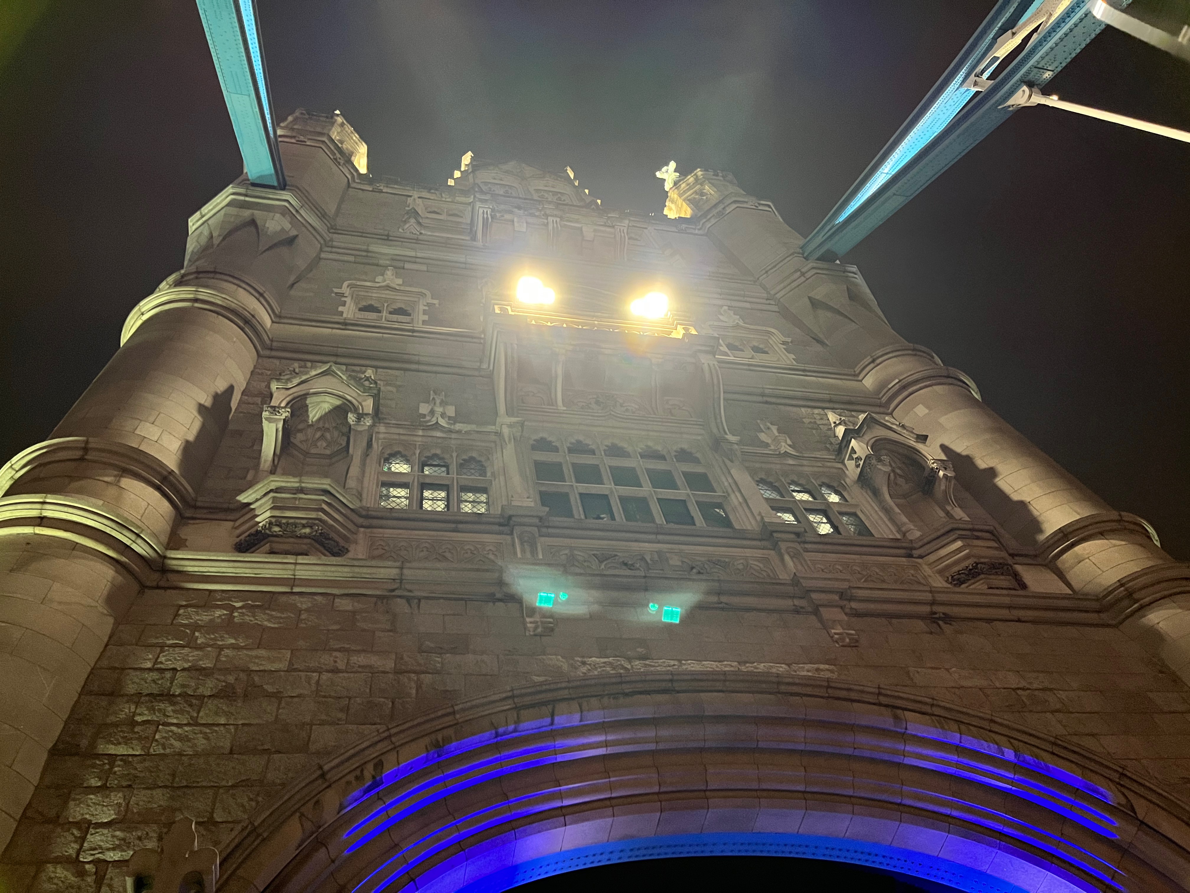 Looking up at one of the ornate stone towers on Tower Bridge. Two bright lights in the centre of the tower are burning down at us, as if it has 2 fiery eyes.