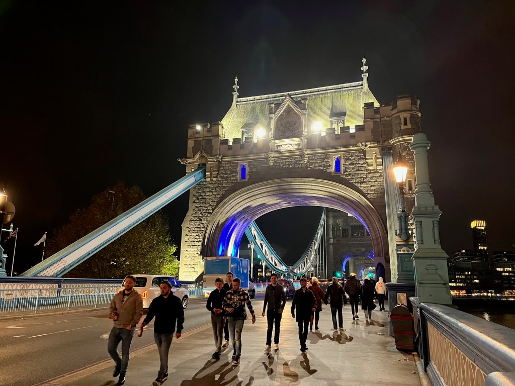 Ornate stone archway across the road at the entrance to Tower Bridge, with blue light on the underside of the arch.