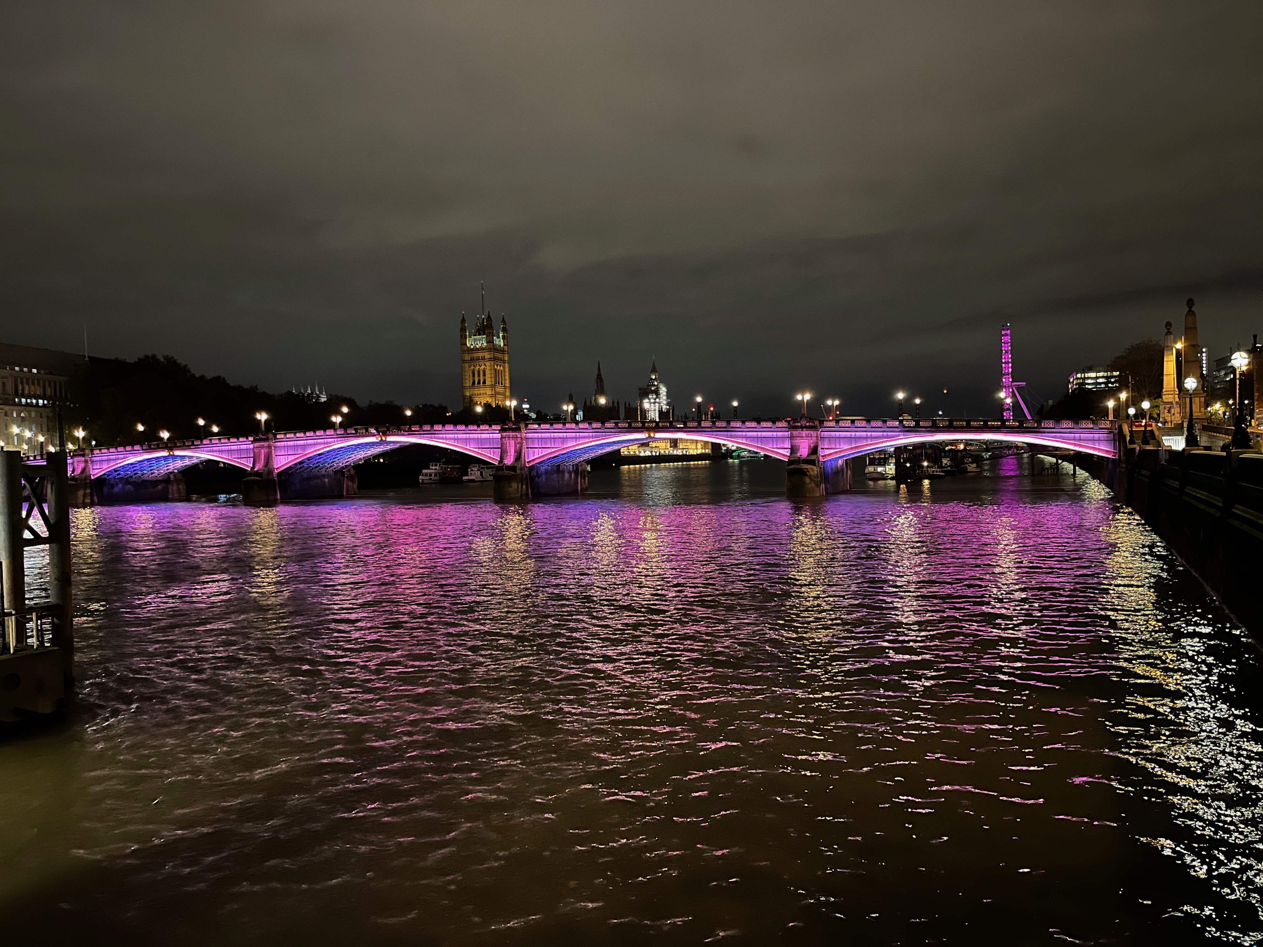Lambeth Bridge bathed in pink light, with blue light visible beneath its 4 arches. The Houses of Parliament and London Eye can be seen on opposite banks of the river in the background.