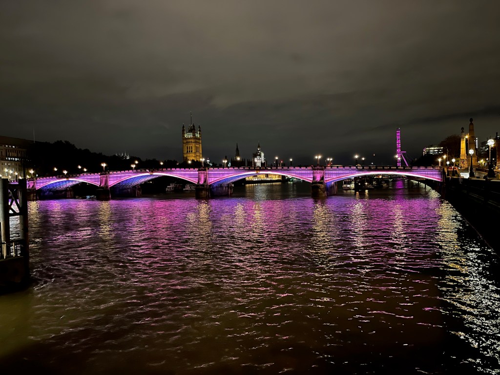 Lambeth Bridge bathed in pink light, with blue light visible beneath its 4 arches. The Houses of Parliament and London Eye can be seen on opposite banks of the river in the background.