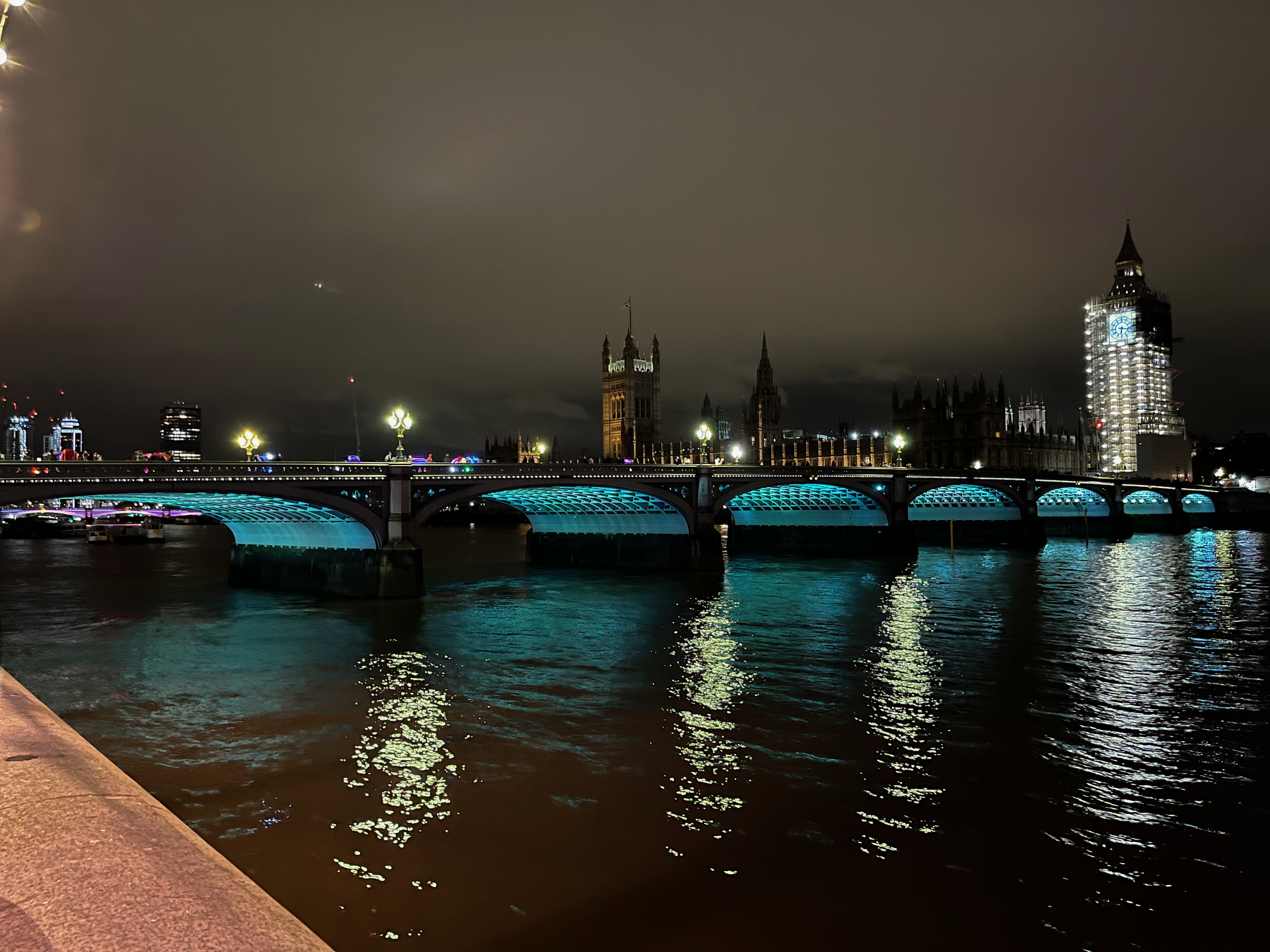 Westminster Bridge, lit beneath its 7 arches in light green. The Houses of Parliament are visible on the opposite bank of the river in the background. Big Ben is covered in scaffolding with a brightly lit clock face at the top.