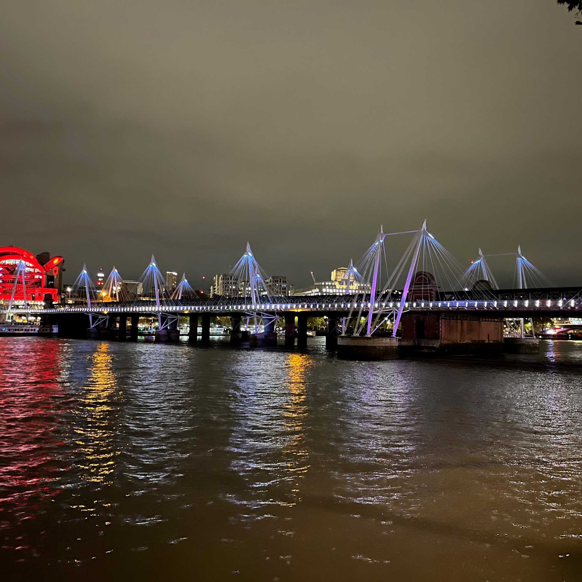 The southern Golden Jubilee Footbridge, a long flat bridge with lots of white lights along the side, and groups of suspension cables at intervals along the bridge bathed in blue and pink light.