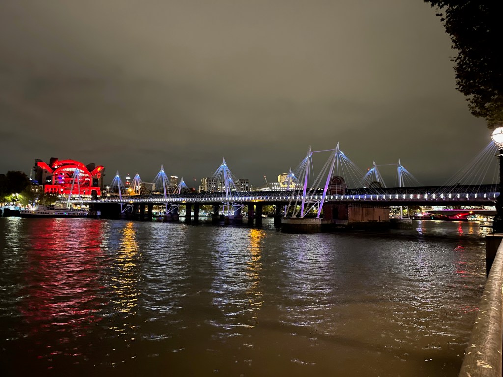 The southern Golden Jubilee Footbridge, a long flat bridge with lots of white lights along the side, and groups of suspension cables at intervals along the bridge bathed in blue and pink light.