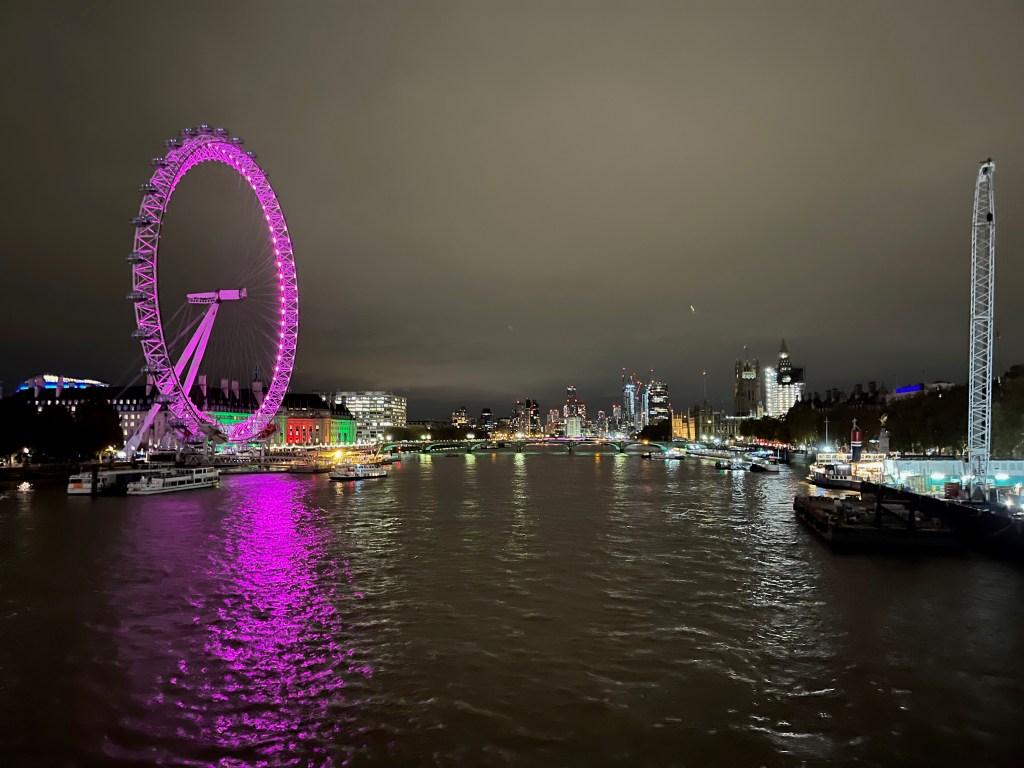 South view of the Thames from the Golden Jubilee footbridge, with the London Eye lit up in pink on the left as it towers high over the buildings behind it.