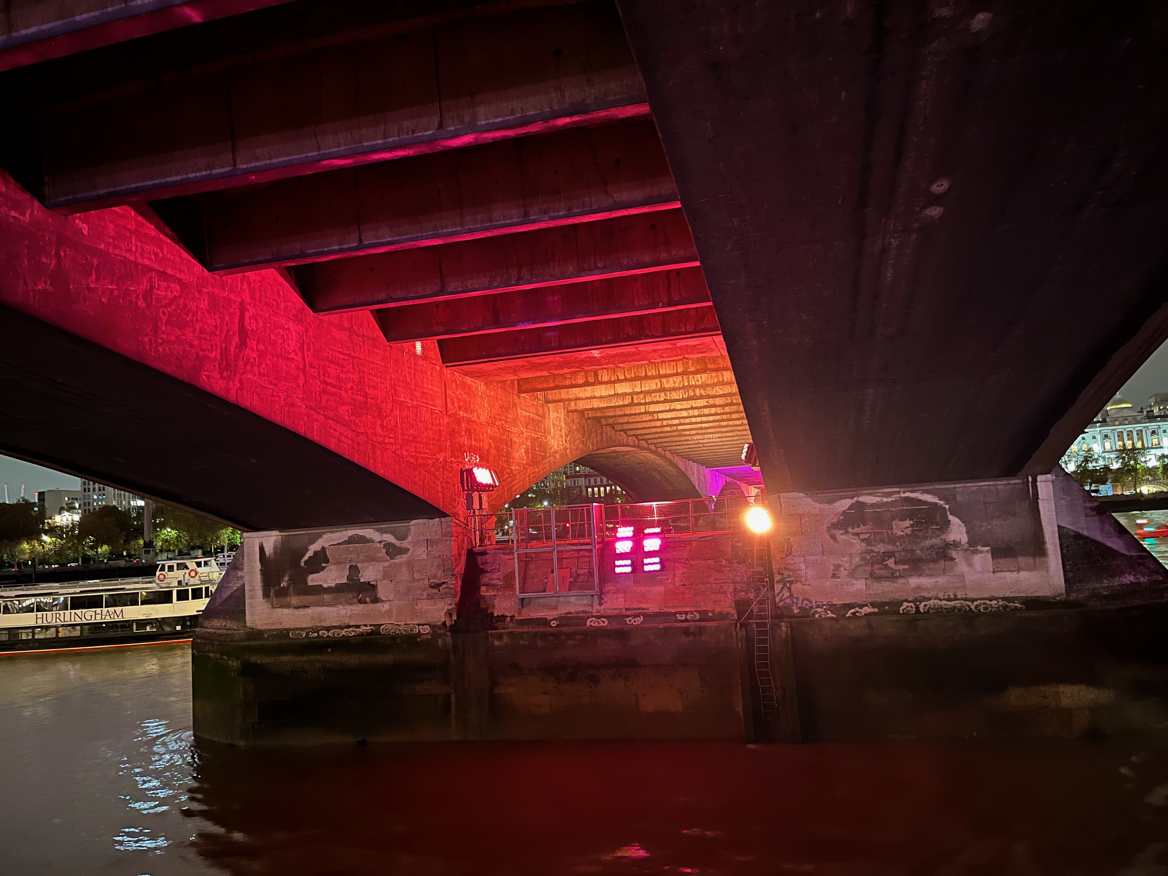 View underneath Waterloo Bridge, where the underside of the bridge is bathed in alternating colours of light including red, orange and pink.