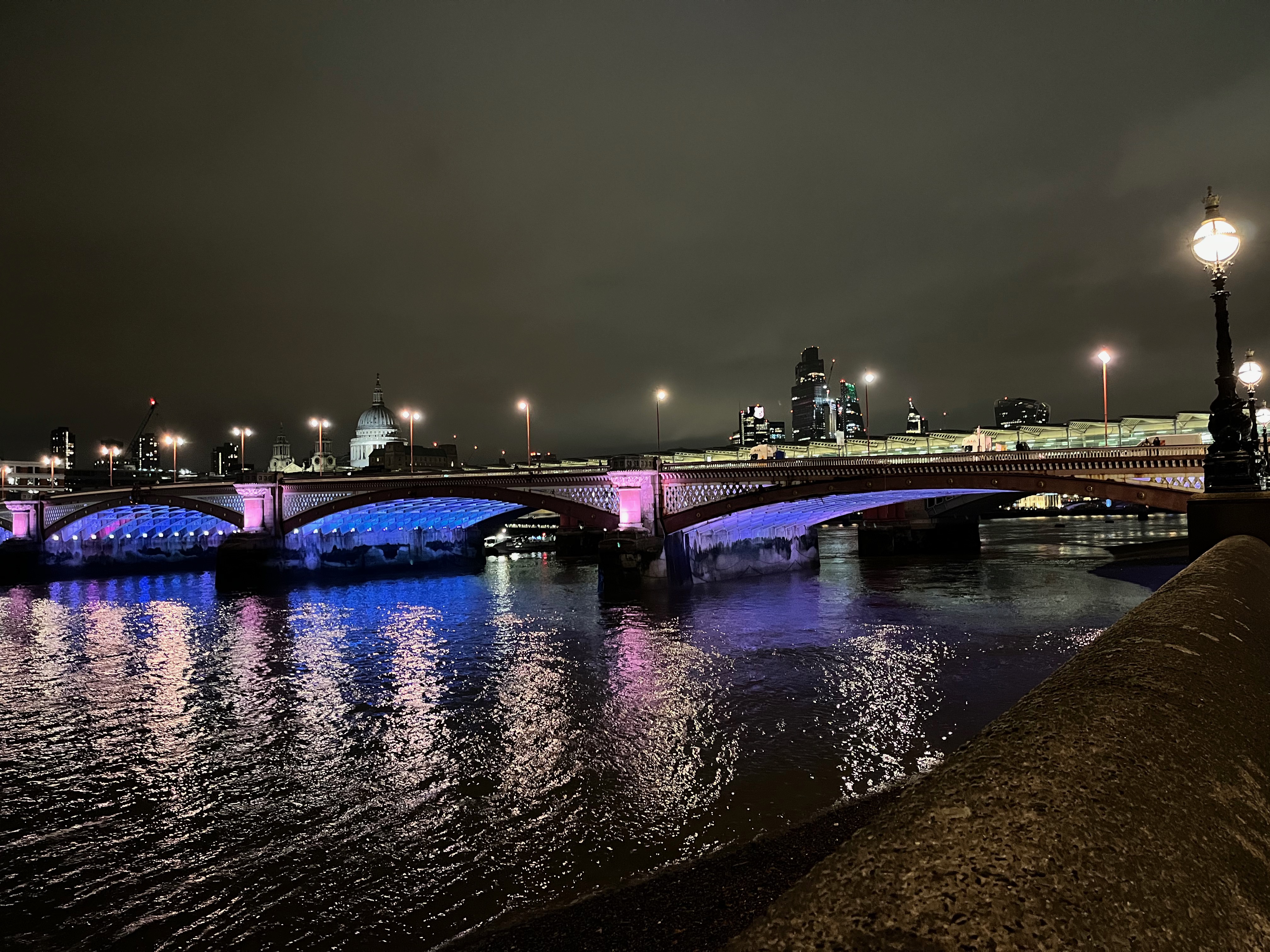 Blackfriars Bridge, with light blue illuminating the underside of its 3 arches. St Paul's Cathedral can be seen in the background.