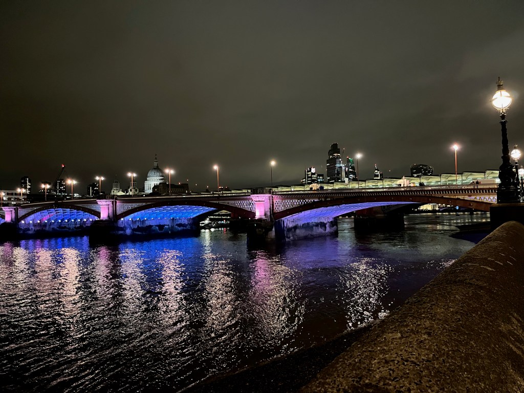 Blackfriars Bridge, with light blue illuminating the underside of its 3 arches. St Paul's Cathedral can be seen in the background.