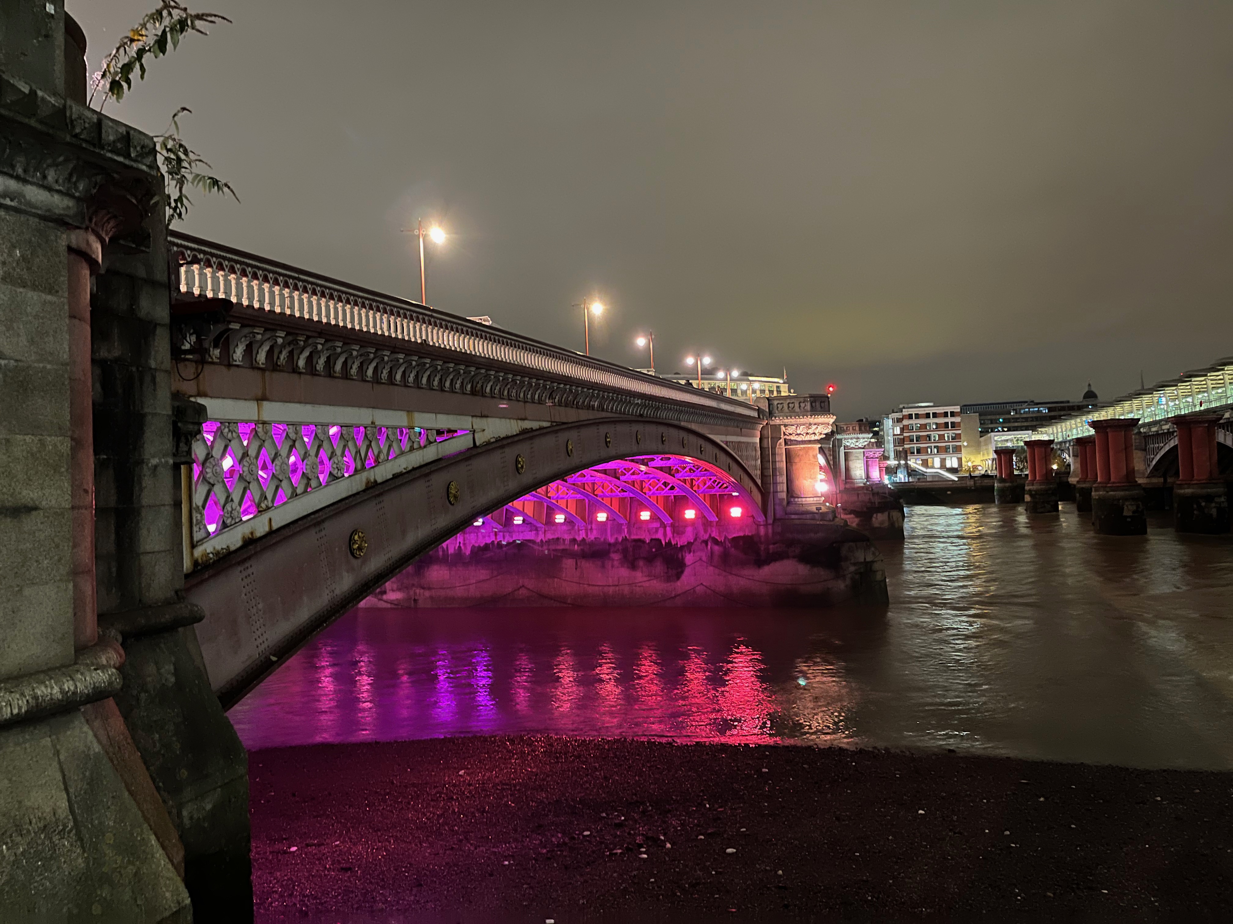 Close-up of one of the arches of Blackfriars Bridge, with the underside bathed in a deep pink light. The Blackfriars Railway Bridge is just visible to the right.