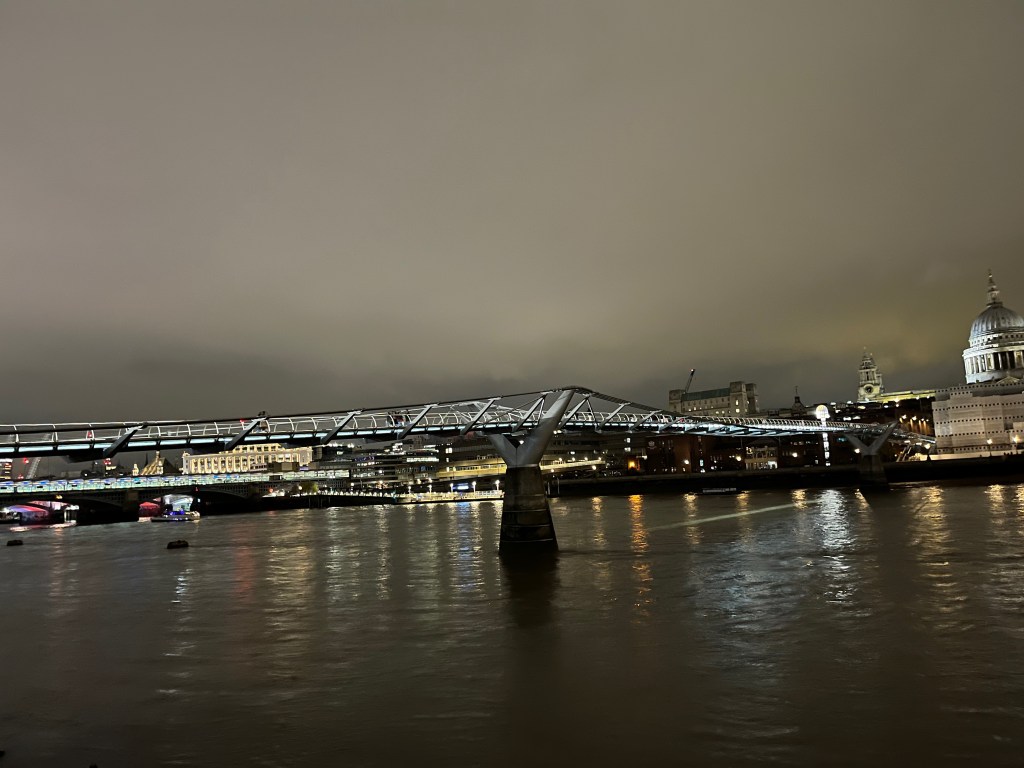 The Millennium Bridge, a suspension bridge that appears to be balanced on a large Y shaped support in he centre of the river, and a similar support on each bank. The edge of the pathway has a white light along the length of the bridge. St Paul's Cathedral can be seen on the far bank.
