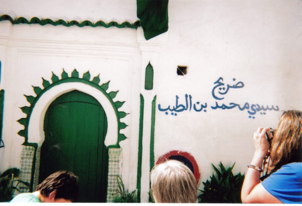 A green doorway in a decorative archway, at the entrance to a large white building in Tangier. There is also Arabic writing on the wall to the right of the door.