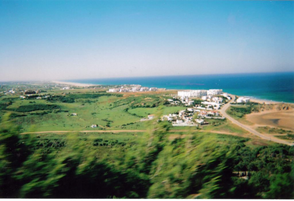 View across a large landscape of green fields and trees, with a cluster of white buildings on the right, and the sea visible in the distance.