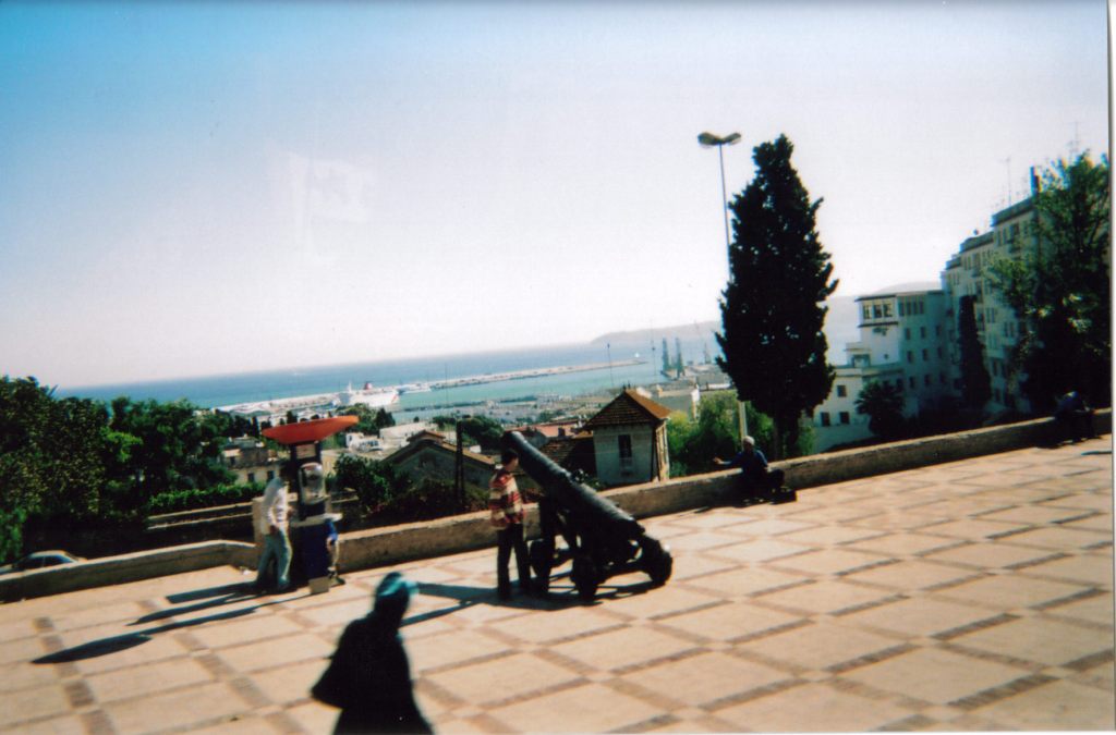 A small black cannon on a paved area is pointing out over the buildings of Tangier below, towards the sea just beyond.