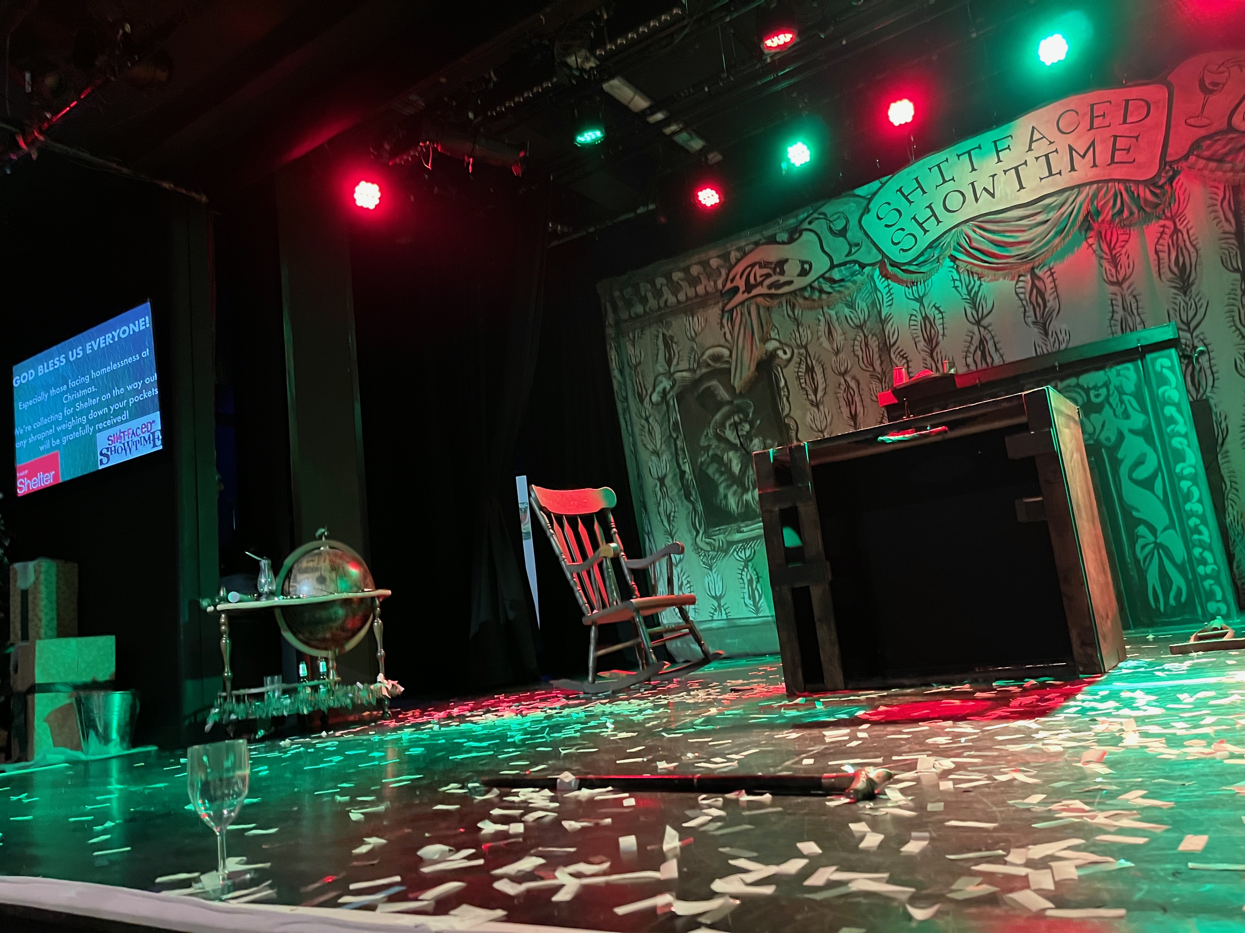 The stage at the end of A Pissedmas Carol, covered in a confetti. A desk and a rocking chair are on the stage, along with a drinks trolley and a bucket to one side. A screen up in the corner of the stage invites people to donate to the Shelter charity to help the homeless.