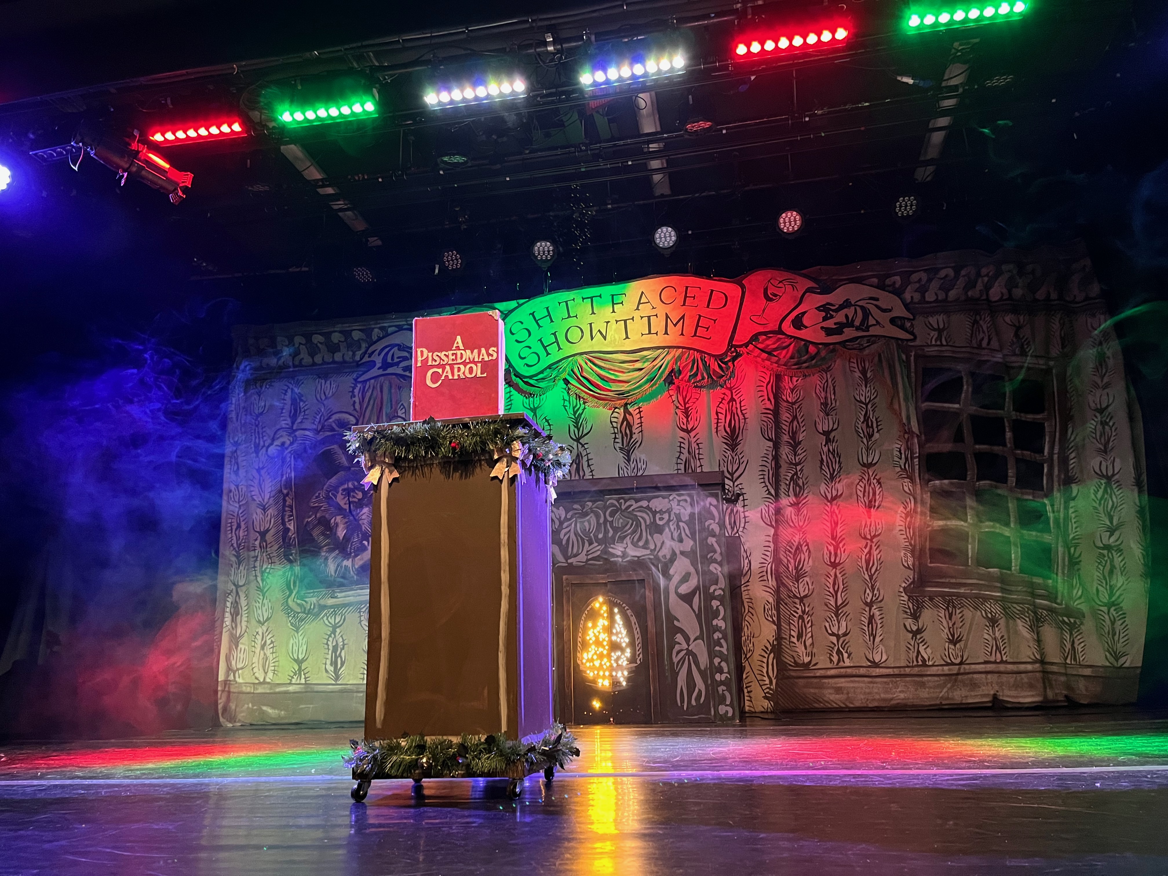 View of the stage for A Pissedmas Carol. A lectern on wheels, decorated top and bottom with tinsel, has a red book on top called A Pissedmas Carol. The backdrop of the stage shows the very decorative walls and fireplace of a Victorian house, with a banner at the top saying Shit-Faced Showtime, bathed in rainbow coloured lighting.