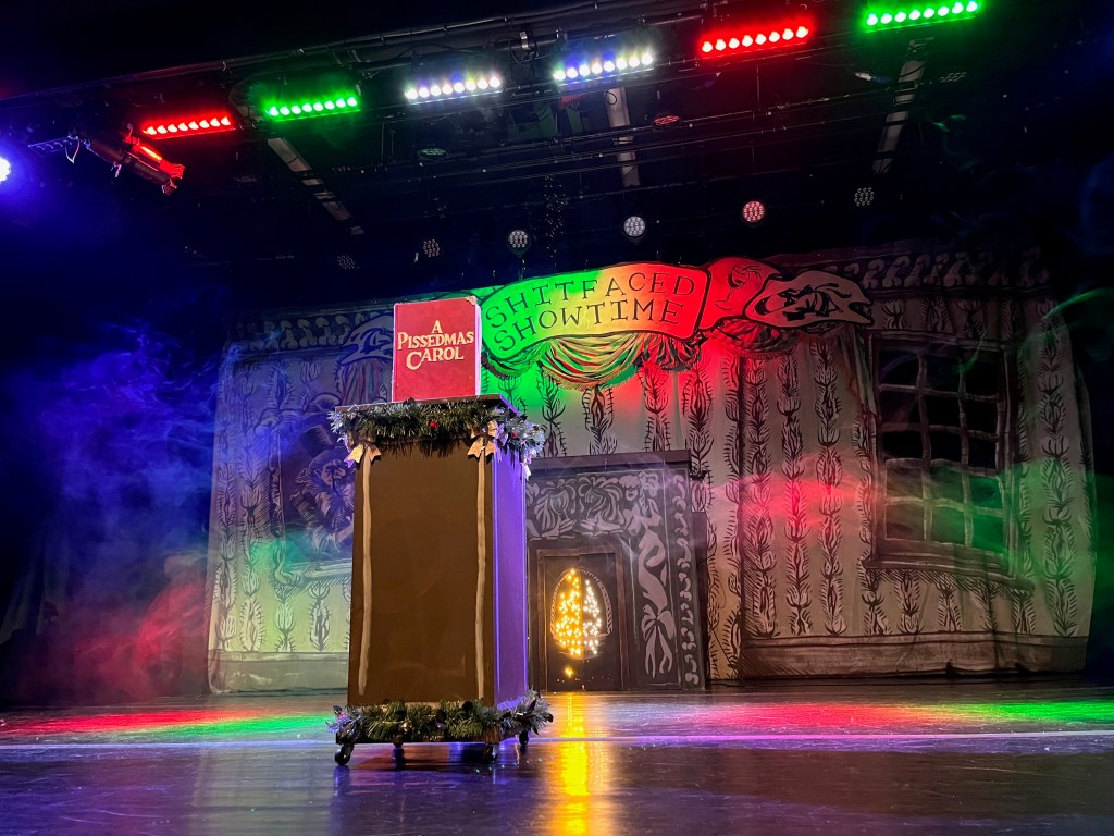 View of the stage for A Pissedmas Carol. A lectern on wheels, decorated top and bottom with tinsel, has a red book on top called A Pissedmas Carol. The backdrop of the stage shows the very decorative walls and fireplace of a Victorian house, with a banner at the top saying Shit-Faced Showtime, bathed in rainbow coloured lighting.