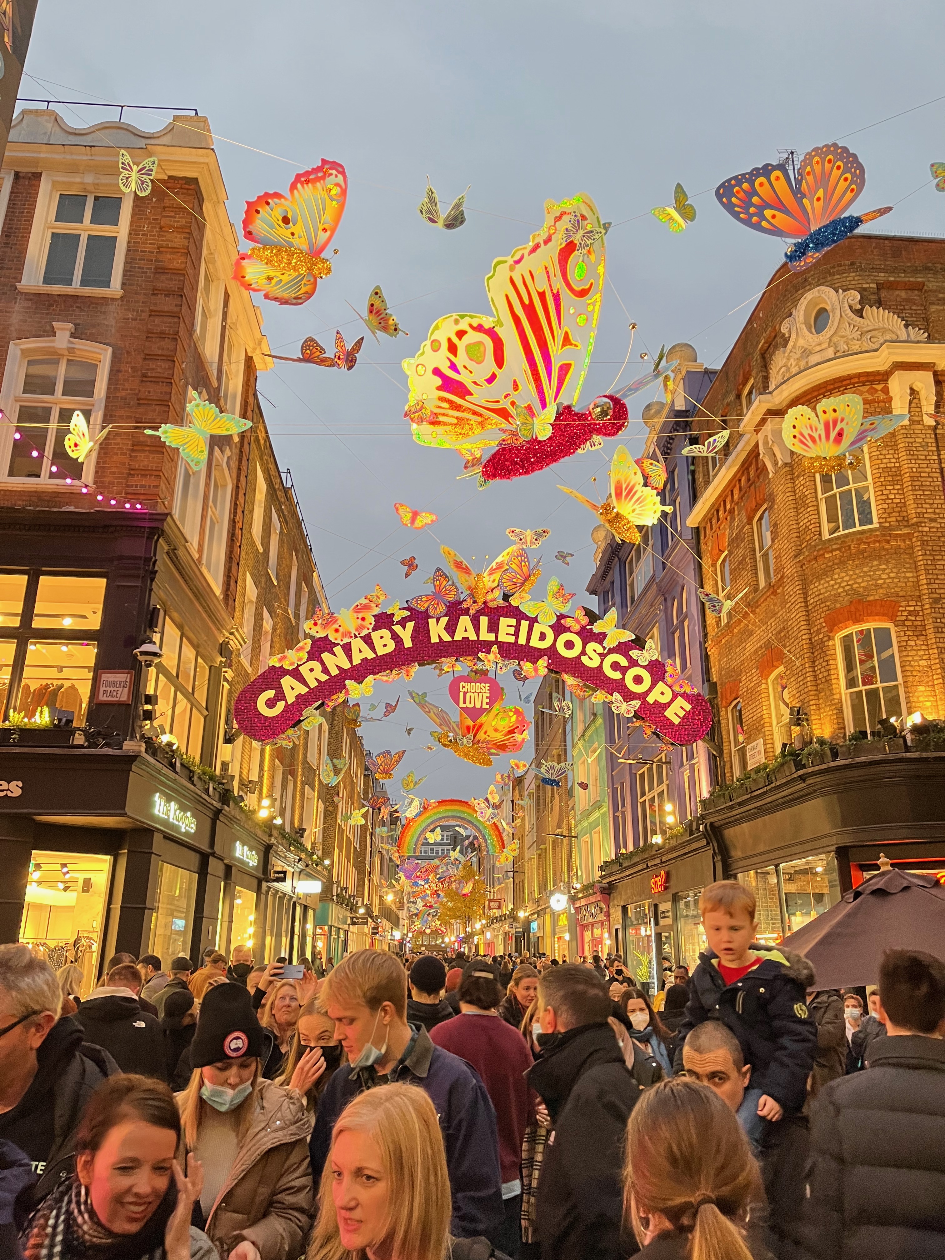 The busy Carnaby shopping street in Central London. In the air, along the length of the street, are lots of colourful, shimmering butterflies of different sizes. In addition, an archway across the street says Carnaby Kaleidoscope with a small heart underneath saying Choose Love, and there's another archway further along designed like a rainbow.