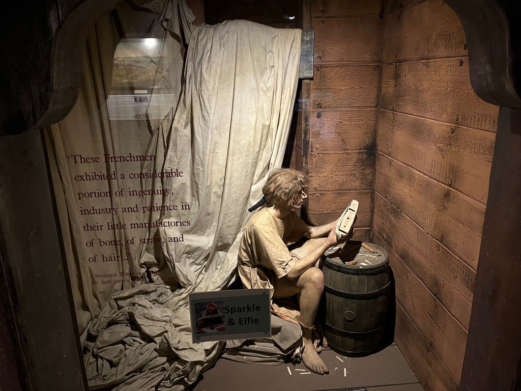 A shabbily dressed prisoner sat in front of a barrel as he works on something in a very small, cramped cell.