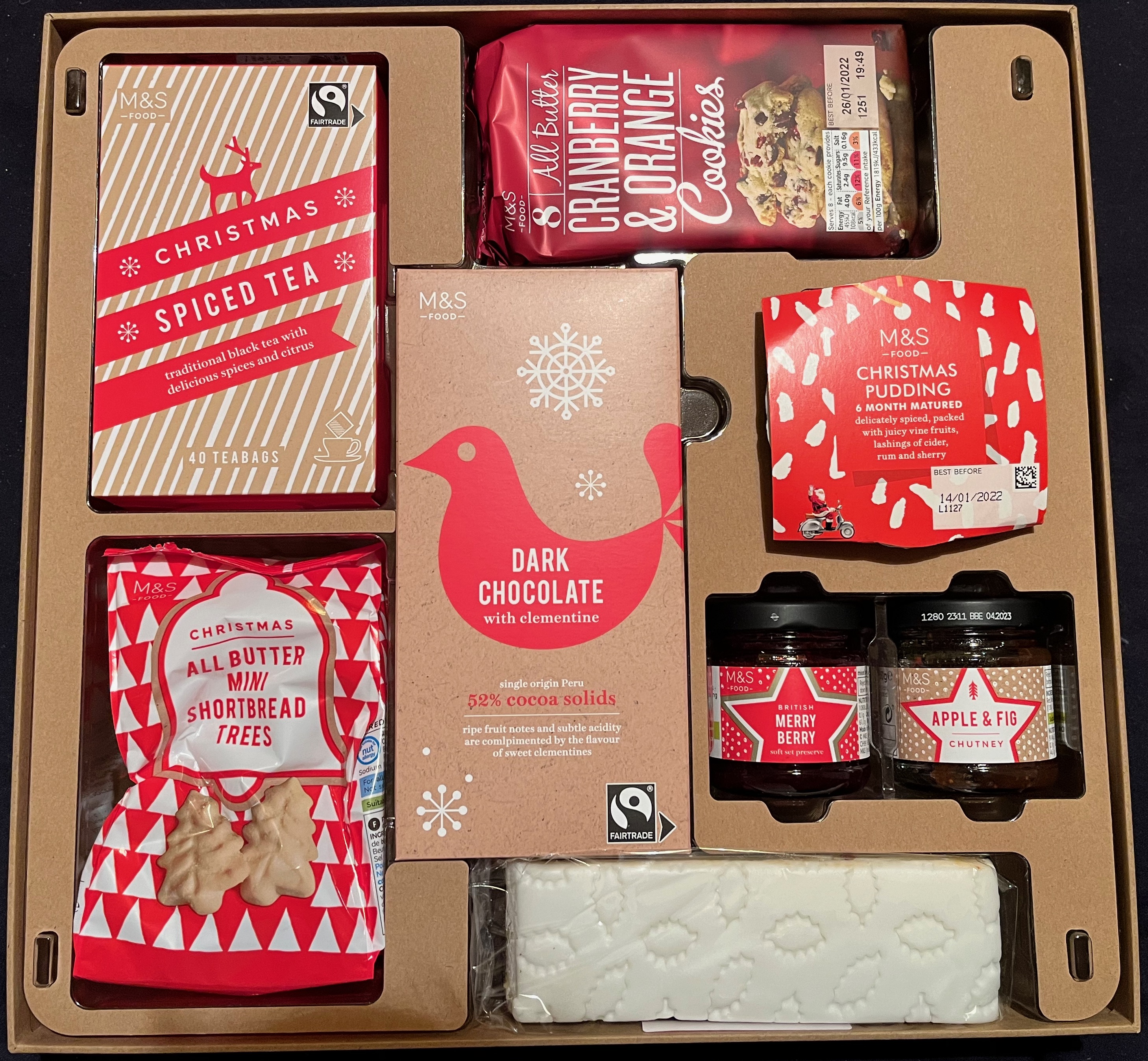 The contents of the Marks And Spencer Classic Hamper, including spiced tea, all butter mini shortbread trees, cranberry and orange cookies, a dark chocolate bar with clementine, a small Christmas pudding, a jar of Merry Berry Preserve, a jar of Apple and Fig Chutney, and a top-iced Christmas cake bar.