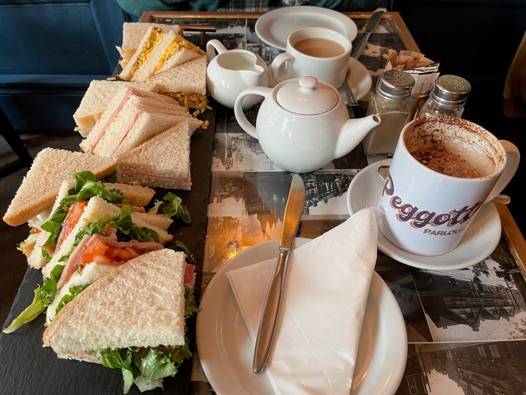Afternoon tea for 2 people from Peggotty's Parlour. One of us is drinking tea, the other hot chocolate. a long plate along one side of the table has 4 BLT sandwiches, 4 ham sandwiches and 4 cheese and pickle sandwiches.