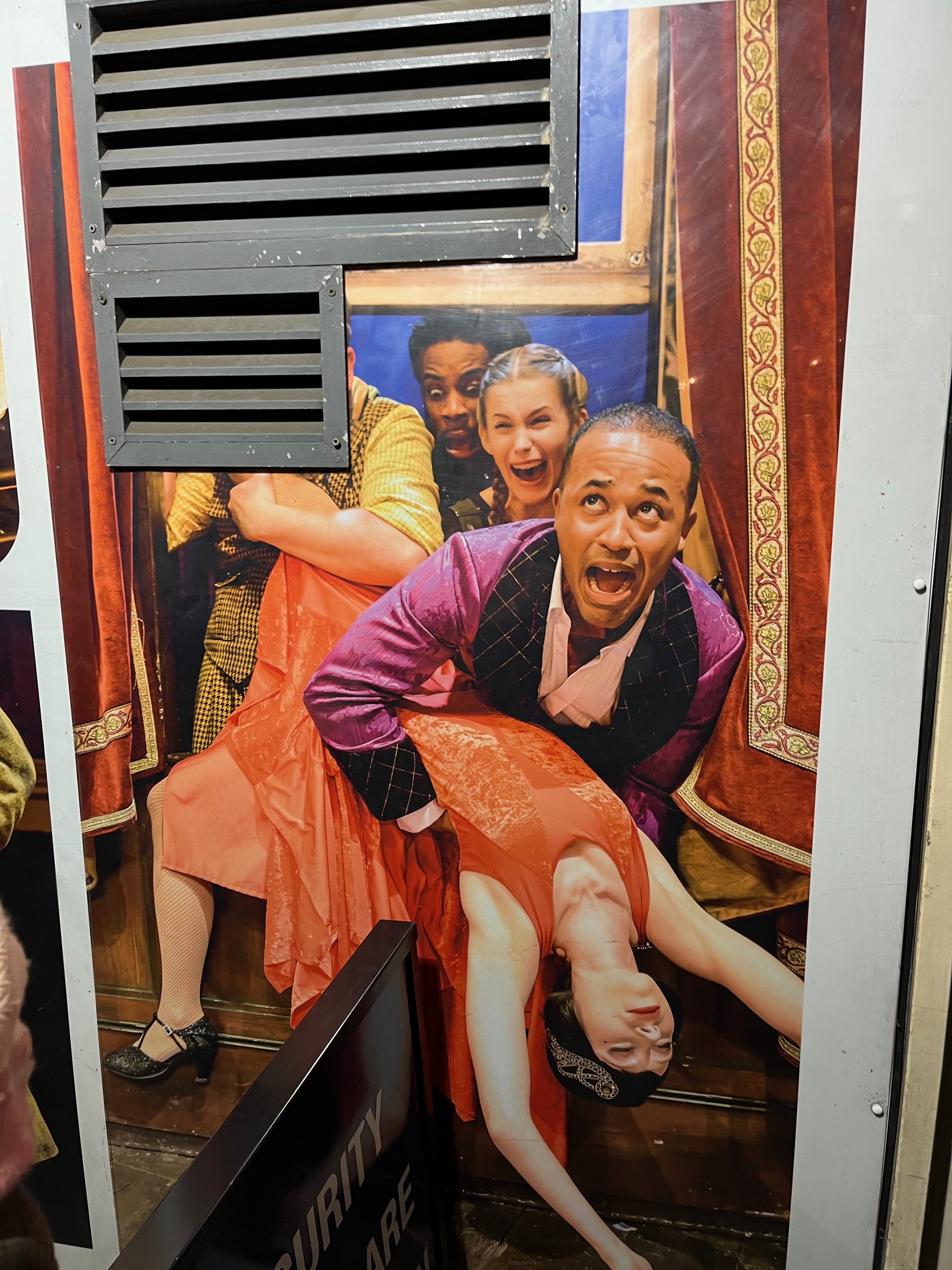 Poster outside the theatre for The Play That Goes Wrong, showing 3 of the characters desperately trying to pull an unconscious lady in a red dress through a window. The poster has been cut to fit around a ventilation grate on the theatre wall, so the head of the character on the left has been cut off.