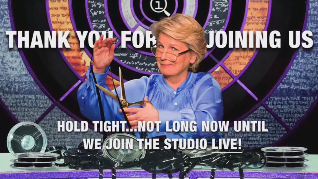 Sandi Toksvig sitting in her hosting chair for QI, with reels of film and tape cluttering the desk. She holds up a reel of tape and prepares to cut it with scissors as she smiles at the camera. Text around her says Thank you for joining us, hold tight, not long now until we join the studio live!