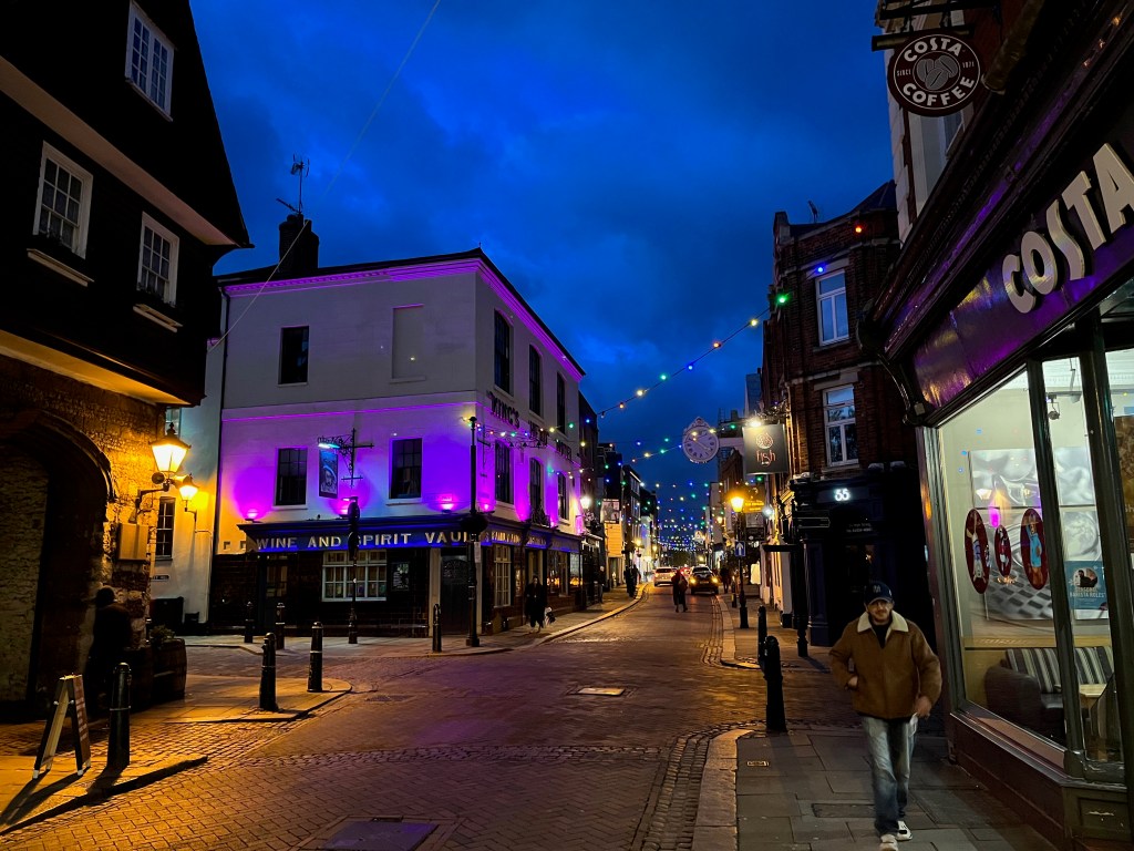The walls of the Kings Head Bar and Hotel lit up in blue and pink, as it stands on the corner of a crossroads. Meanwhile, stretching into the distance along the high street are zig-zagging strings with lots of colourful lights, suspended up high between the buildings on each side.