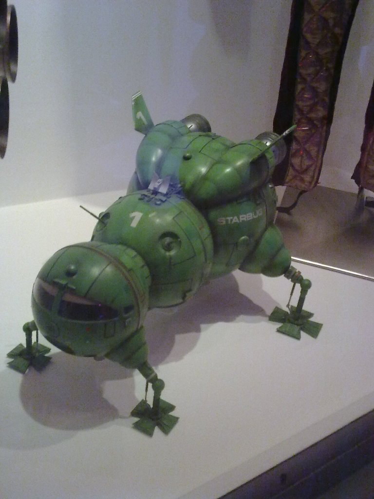 Model of Starbug from Red Dwarf, a small space ship made of 3 spherical sections that increase in size from front to back. Thin legs with cross-shaped feet stick out at right angles from the 4 corners of the model to hold it up.