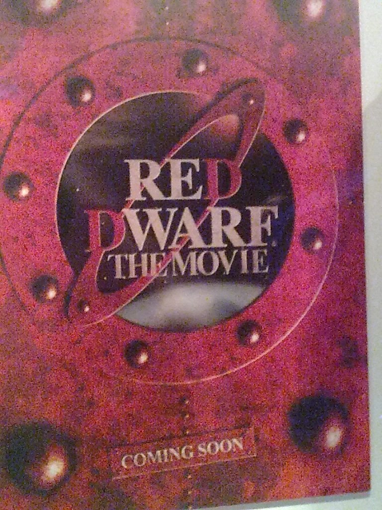 Teaser poster for Red Dwarf The Movie, with the title placed over a round porthole window of the red spaceship, looking out into space. Text on the wall below the window says Coming Soon, but the movie never got made in the end.