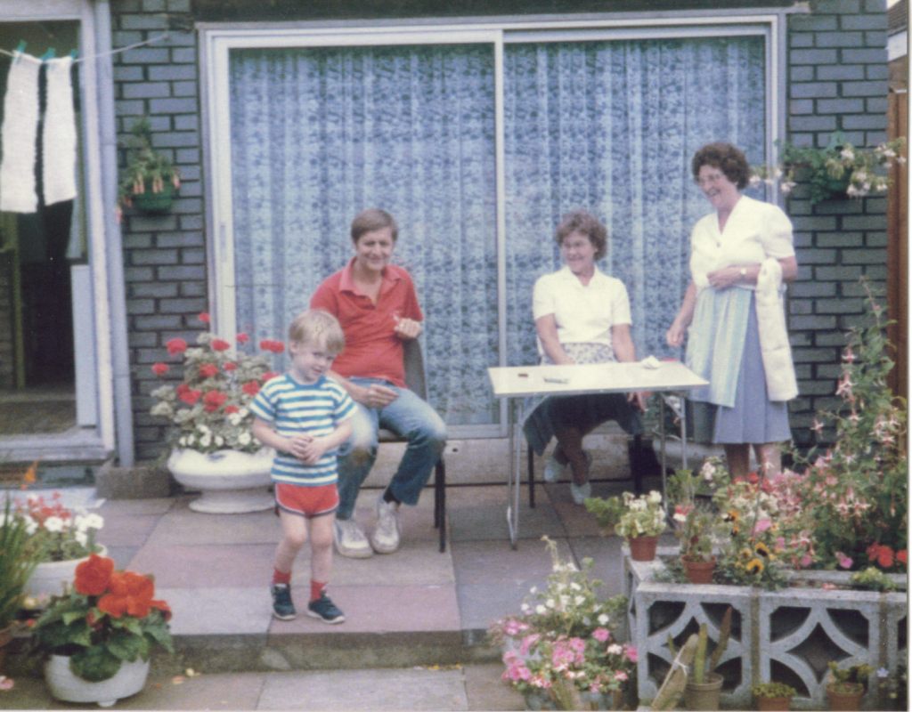 Glen, as a young child, is standing and smiling on a paved garden patio, wearing a blue and white striped t-shirt, red shorts and black trainers. His father, sat behind him in a red shirt, blue jeans and white trainers, watches with a smile, alongside 2 female relatives. Flower pots nearby contain a mix of red and white flowers.