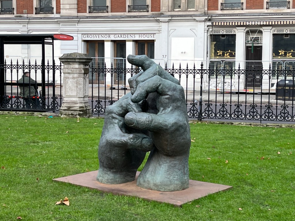 A sculpture of 2 large hands that appear to be reaching out of the ground and touching each other with their fingers. The sculpture is in a public garden, surrounded by grass.