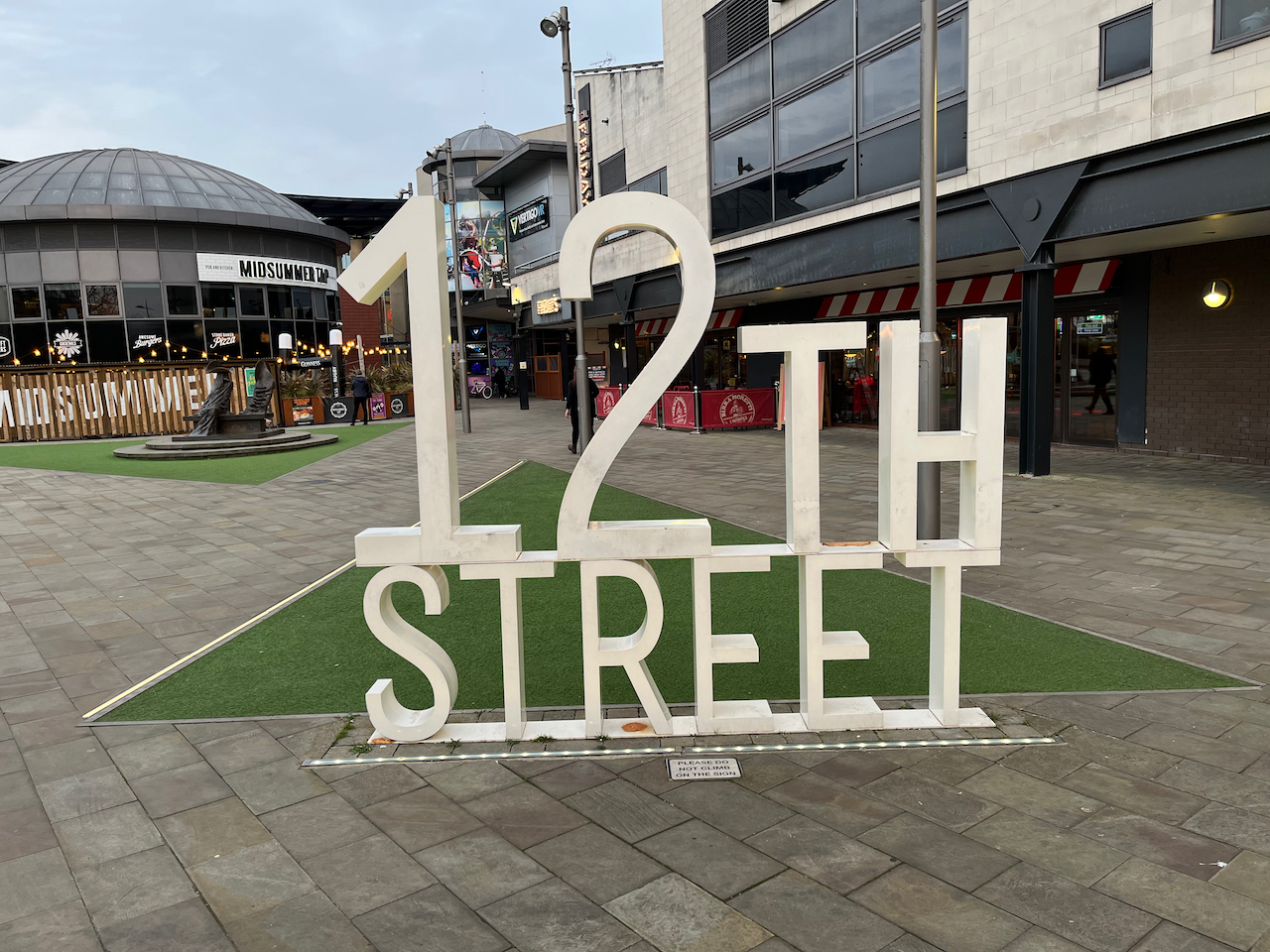 A free-standing 3D sign of large white text, with 12th on the top and the word Street underneath it. There's a pub and restaurants in the background.