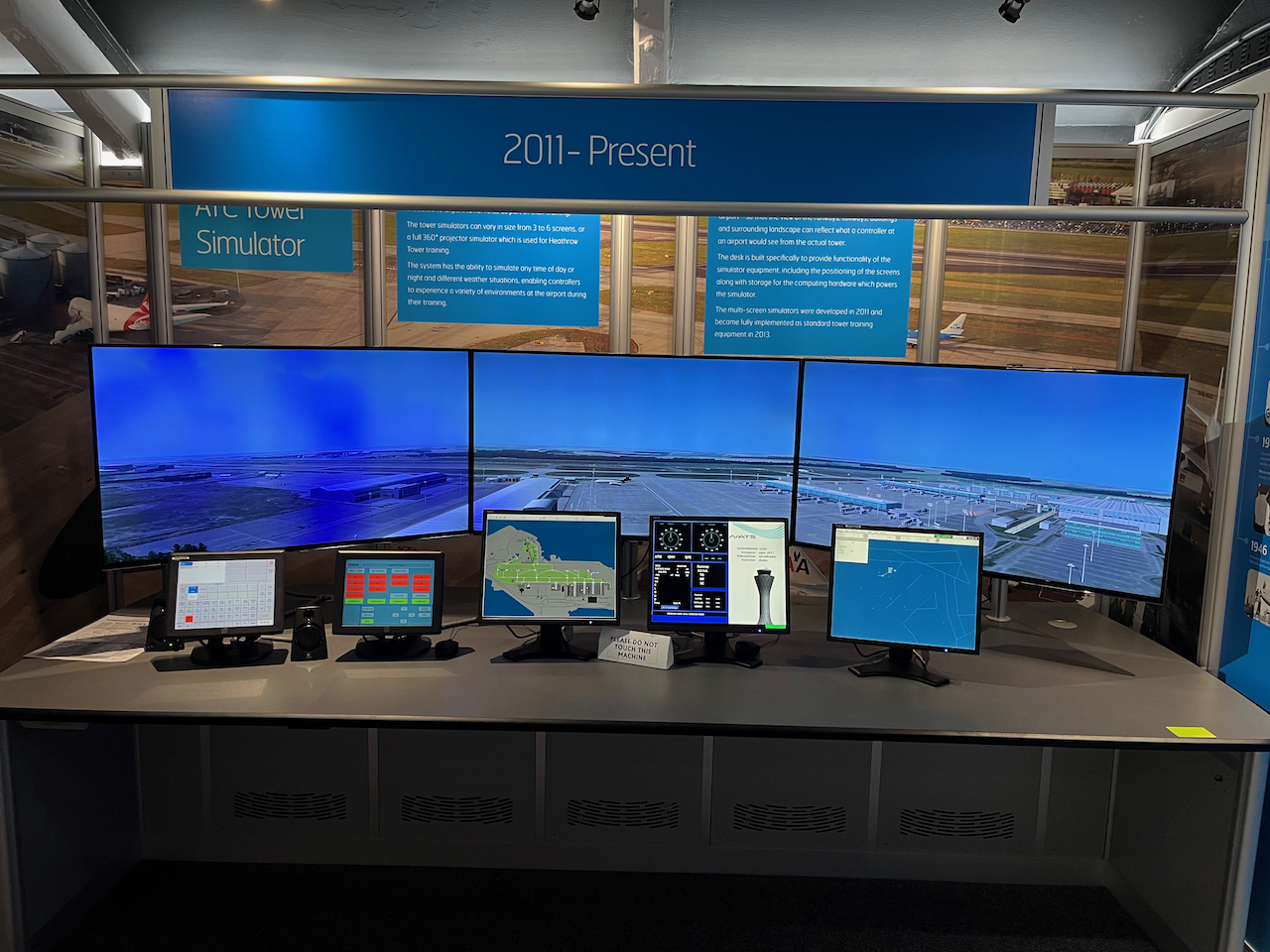Air traffic control tower simulator. A single large image of an airfield is spread across 3 large monitors, to represent looking out of the tower window. 5 smaller screens on the desk in front are displaying lots of different information.