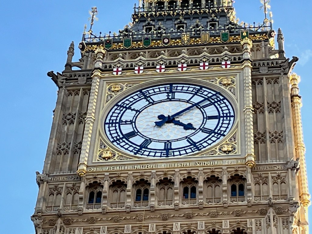 Close-up of Big Ben's black and white clock face, framed by the ornate golden architecture surrounding it, including 5 tiny shields across the top featuring a red cross on a white background.