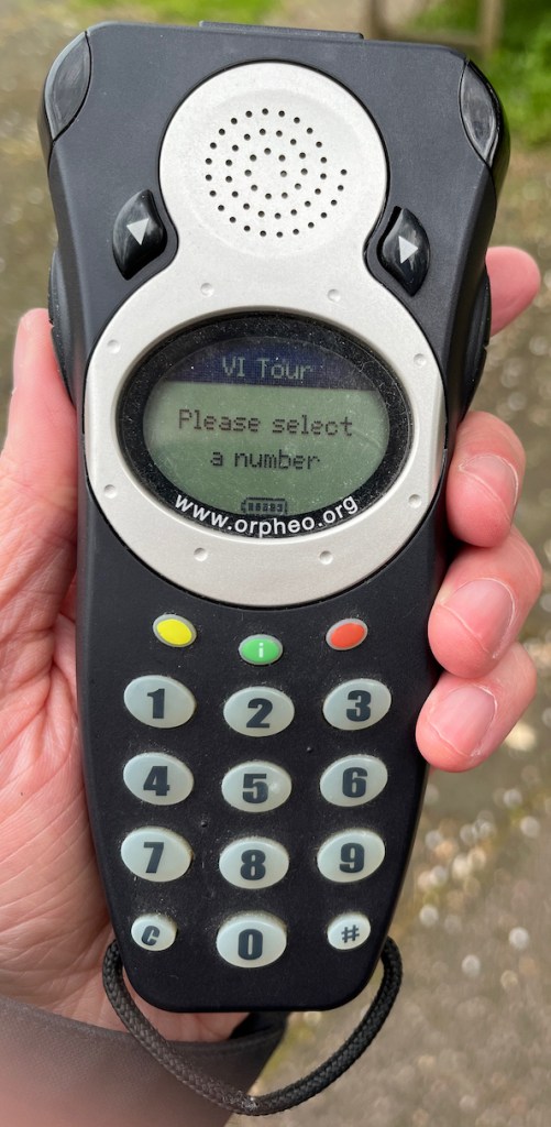 Audio description handset. From the top down, there is a speaker with left and right arrow buttons on each side of it, then a round screen with the text VI Tour, please select a number, then 3 coloured keys in yellow, green and red, then a numeric keypad.