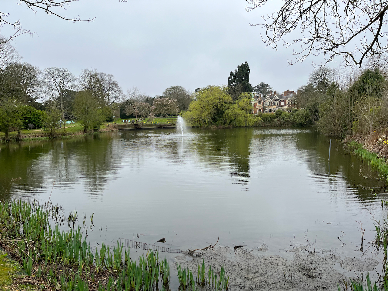 The big lake in Bletchley Park, surrounded by trees and grassy areas, with a fountain near the far bank.