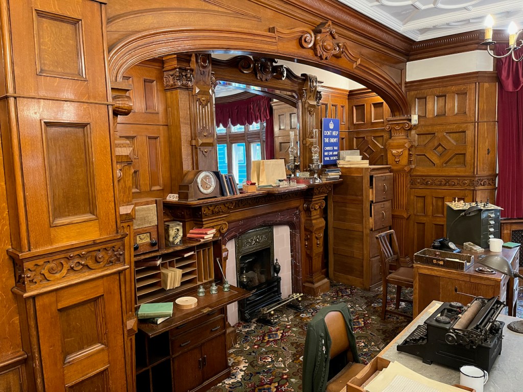 The ornate fireplace in Bletchley Park library, above which is a large mirror. The mantlepiece holds a clock, pieces of paper and other items. A table to the left of the fireplace holds a small document organiser with several pigeon holes and a few books sticking out of them, and on top of it is a radio. A blue poster to the right of the fireplace says Don't help the enemy, careless talk may give away vital secrets.