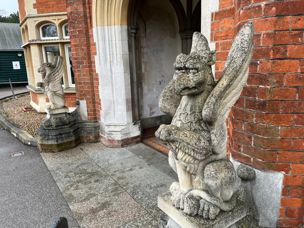Stone gargoyles on either side of the entrance to the Bletchley Park mansion. They are large scaly-skinned creatures sitting on their hind legs, with large wings sticking out behind them.