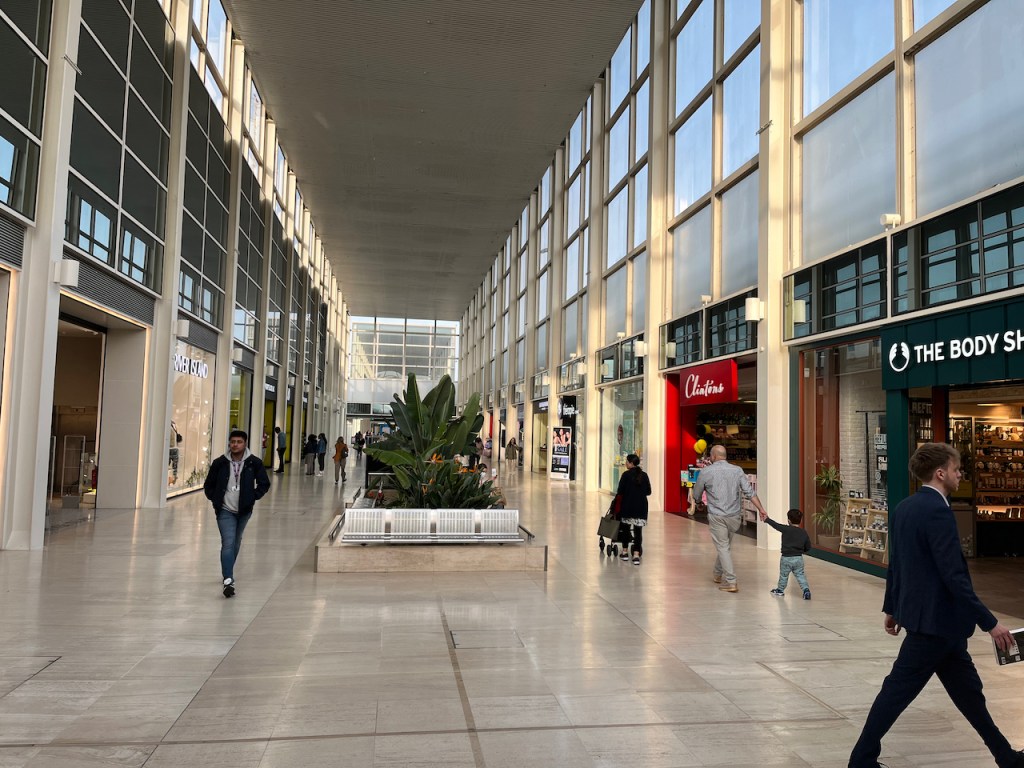 Long corridor of shops in the Centre MK shopping centre, including The Body Shop, Clinton's Cards and River Island. In the middle of the wide walkway is a metal bench in front of some plants with big green leaves.