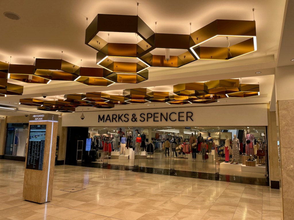 Wide entrance to Marks & Spencer at the end of the Centre MK corridor. A pattern of big bronze hexagons are suspended from short wires on the ceiling in front of the entrance.