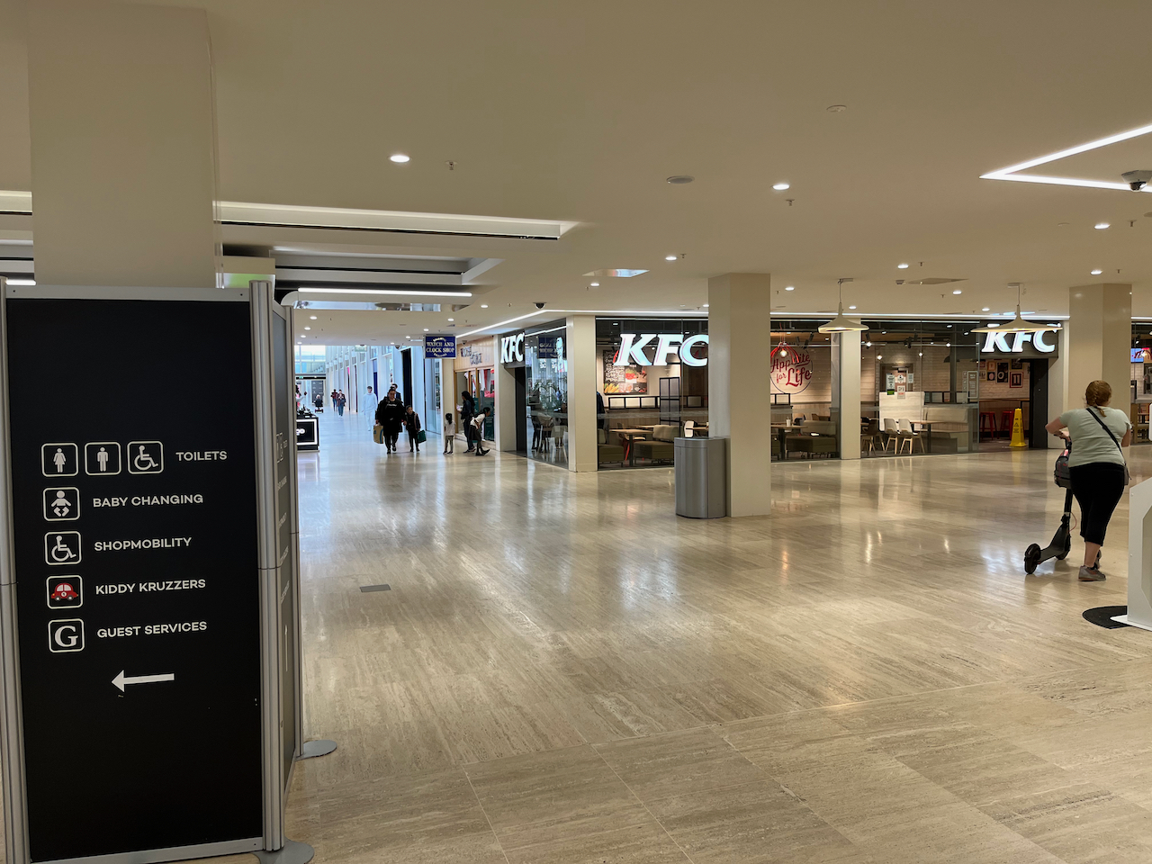 View towards KFC on the corner of a side corridor in Centre MK. A tall blue sign states that turning left leads to the toilets, baby changing, shop mobility, kiddy cruisers and guest services.