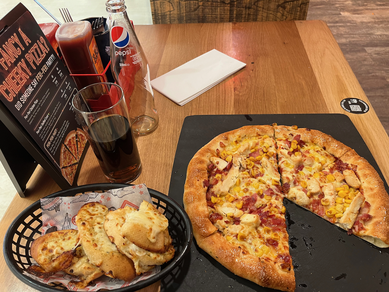 Meal in Pizza Hut, consisting of an 11-inch BBQ Americano Stuffed Crust pizza, some gralic bread with Cheese, and a small bottle of Pepsi. A small round black sticker on the table shows the table number, and the QR code you scan to place your order.
