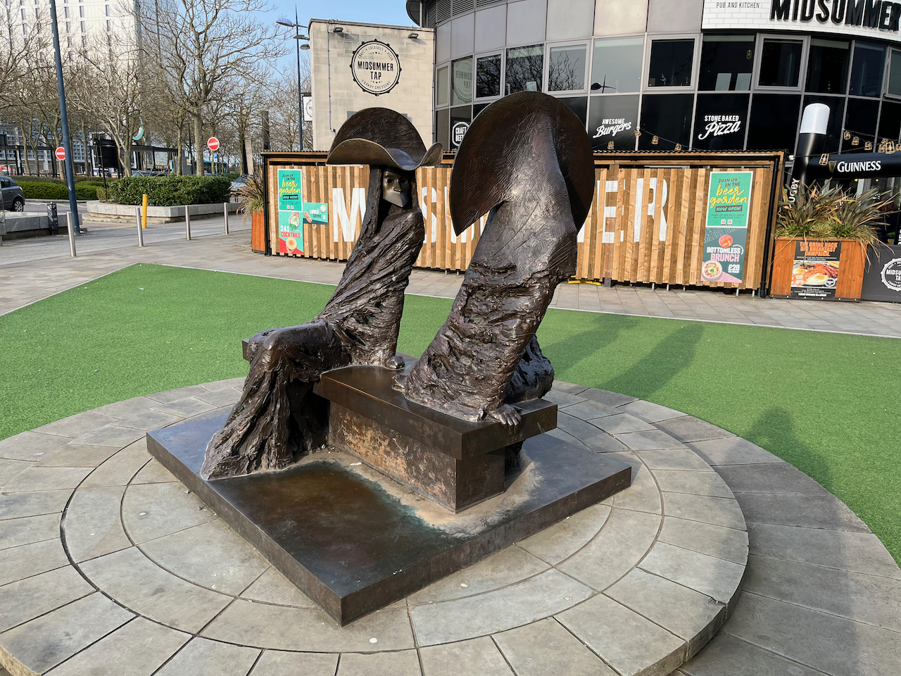 A sculpture called Dangerous Liaisons by Philip Jackson, showing 2 figures sitting on a bench next to each other, facing in opposite directions. Both figures are wearing long flowing capes, full face masks with a sharply pointed nose and chin, and large hats with a big brim sticking up at the back and a wavy brim at the front.