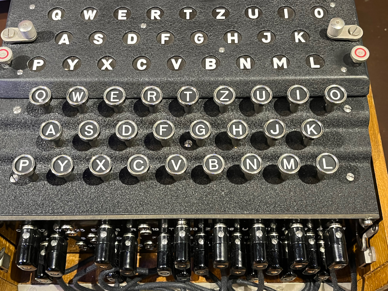 Close-up of the Enigma keyboard. It uses the German QWERTZ layout which, compared to the QWERTY keyboard we're used to, swaps the Z and Y keys around, then moves the P and L keys to the far left and right of the bottom row. The same layout of letters is replicated in circular lights embedded above the keyboard. Whenever a key is pressed, a different letter is lit up above. It's never the same as the letter that's been pressed, and pressing the same key multiple times causes a different letter to light up every time.