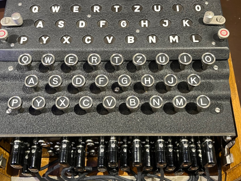 Close-up of the Enigma keyboard. It uses the German QWERTZ layout which, compared to the QWERTY keyboard we're used to, swaps the Z and Y keys around, then moves the P and L keys to the far left and right of the bottom row. The same layout of letters is replicated in circular lights embedded above the keyboard. Whenever a key is pressed, a different letter is lit up above. It's never the same as the letter that's been pressed, and pressing the same key multiple times causes a different letter to light up every time.