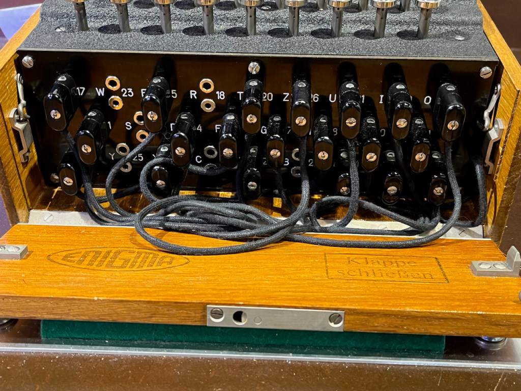 Close-up of the plugboard on the front of the Enigma machine. There are 26 pairs of headphone-style sockets, set in the German QWERTZ keyboard layout, and each pair of sockets is arranged vertically. The letter of the alphabet is written to the left of each pair, and the corresponding number to the right e.g. D 4, R 18 and Z 26. Wires are plugged into several of the sockets.