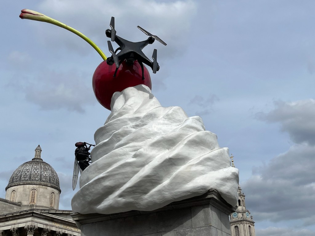 Artwork on the Fourth Plinth in Trafalgar Square. The main sculpture is a swirling tower of white ice cream, like you get in an ice cream cone, topped by a large red cherry with a very tall green stem. A large black fly sits on the back of the ice cream, while a large black drone is attached to the front of the cherry.