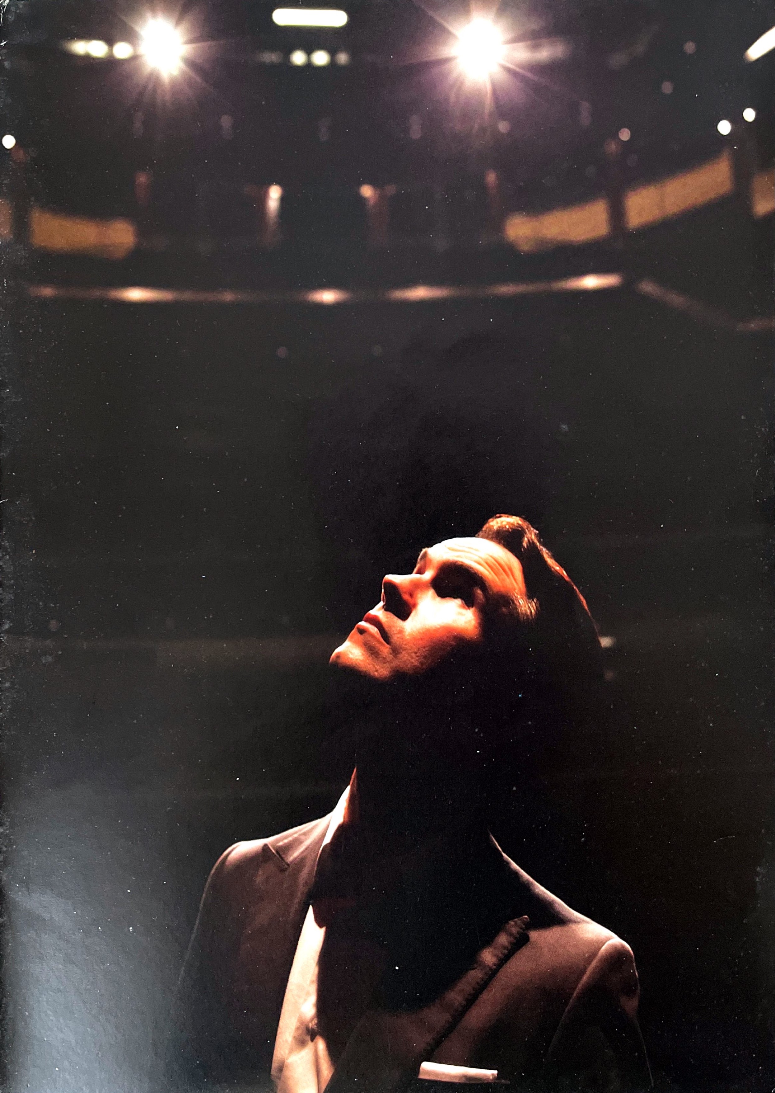 A photo of Jimmy Carr looking upwards thoughtfully, as a spotlight illuminates his face in a dark theatre.