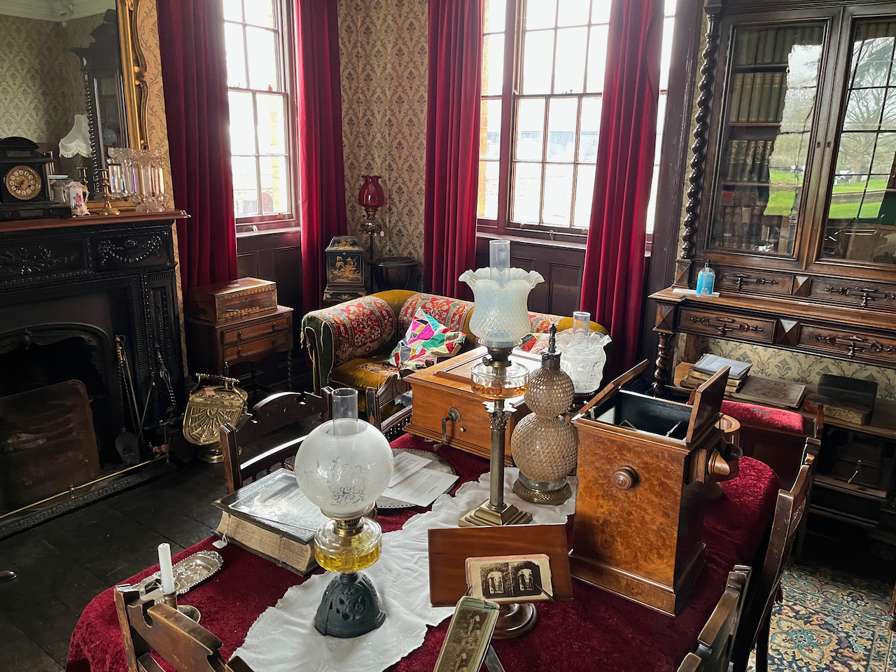 An ornately decorated and lavishly furnished parlour room, including a fireplace and a sofa, and a table containing many items including a few different types of music boxes and lamps.