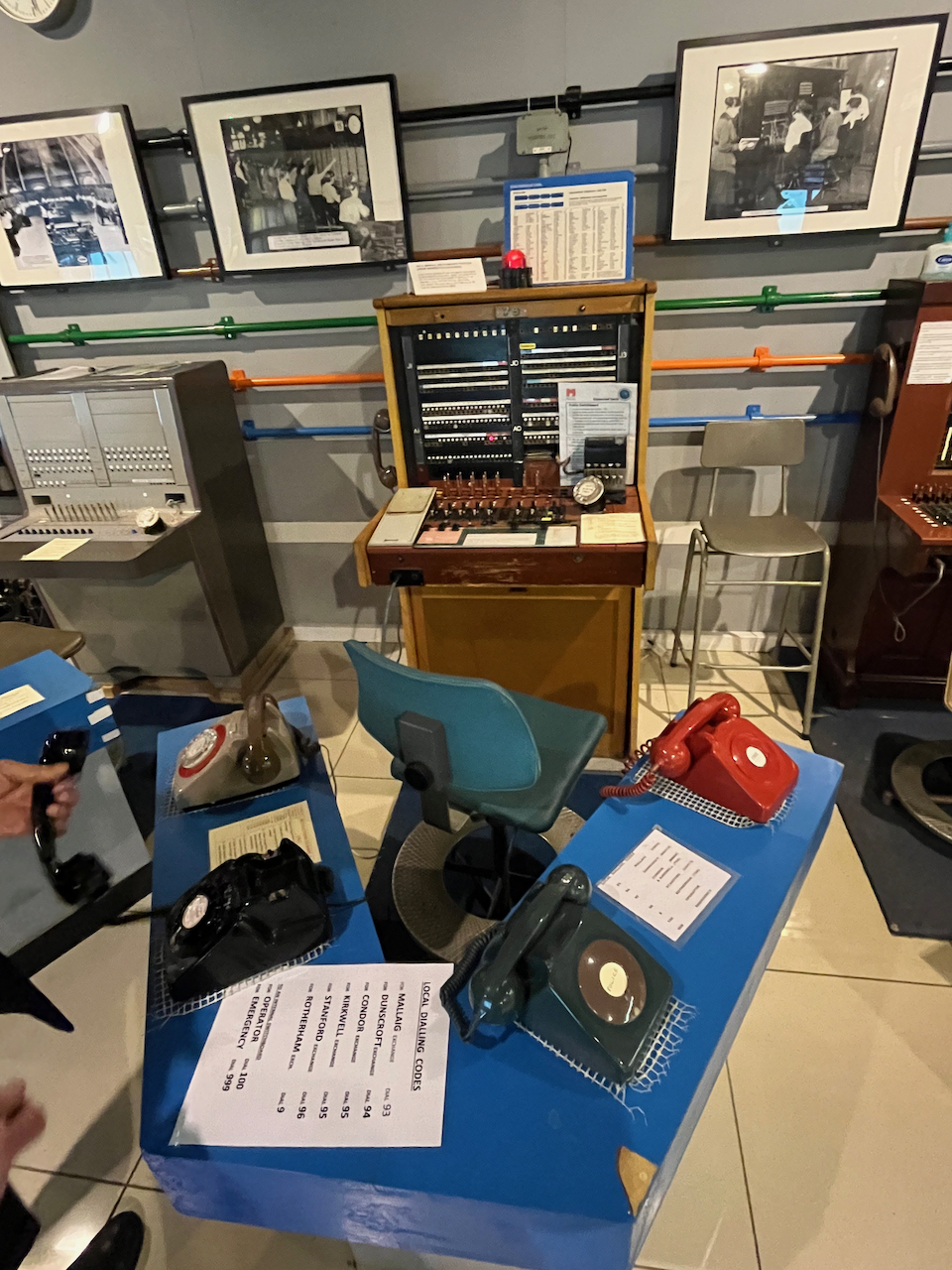 A desk with 4 telephones in front of a telephone exchange unit on another desk, which has lots of sockets and controls.